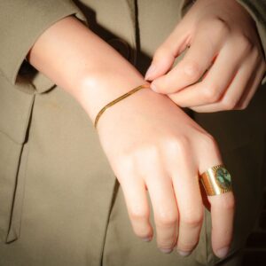 Bracelet plat doré (Venn) porté au poignet droit, tenu avec la main gauche. Une bague au doigt