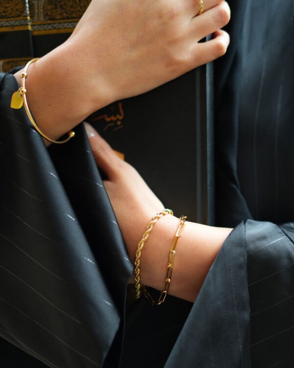 bracelet rigide, doré, avec un coeur à personnaliser, sur un poignet. L'autre poignet possède deux bracelets doré différents